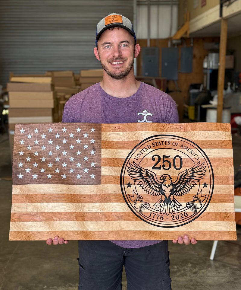 Man holding a wooden sign with American flag design and '250' engraving in a workshop.