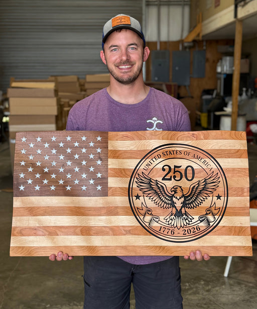 Man holding a wooden sign with American flag design and '250' engraving in a workshop.